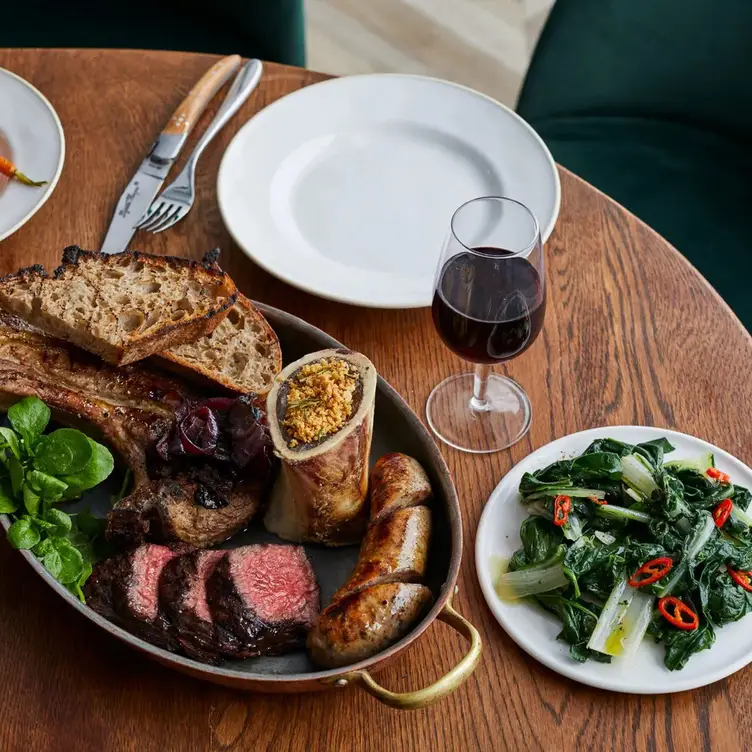 A meat platter featuring sliced medium-rare steak, bone marrow, sausage, grilled bread, and watercress, alongside a glass of red wine and a side of sautéed greens with chilli
